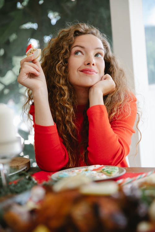 From below of dreamy female with decorated cookie in hand looking up while sitting at festive table with various food during Christmas holiday