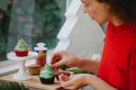 Side view of female cook putting sprinkles on frosting of cupcake while standing near window during holiday preparation at home