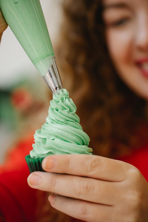 Crop blurred female confectioner with curly hair in red outfit decorating yummy homemade cupcake with green whipped cream