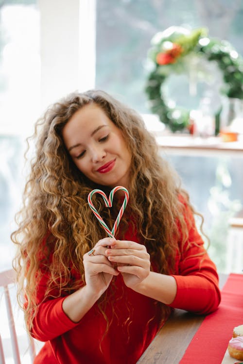 Happy long haired curly woman in red dress showing candy canes in form of heart in studio with Christmas decorations at daytime