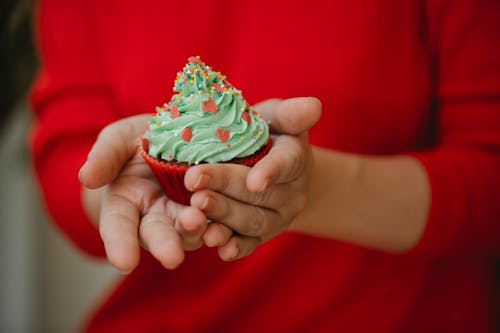 Crop anonymous lady in red dress demonstrating delicious cupcake decorated green whipped cream and colorful sprinkle on kitchen