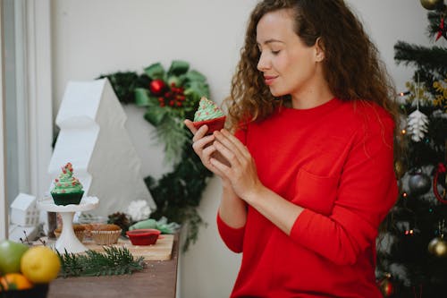 Happy young woman enjoying fresh delicious cupcake with green whipped cream standing beside decorated Christmas fir tree