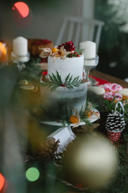 Free Christmas table with white tasty cake decorated with physalis and garnet near candles and leaves near branches with cones in light room Stock Photo