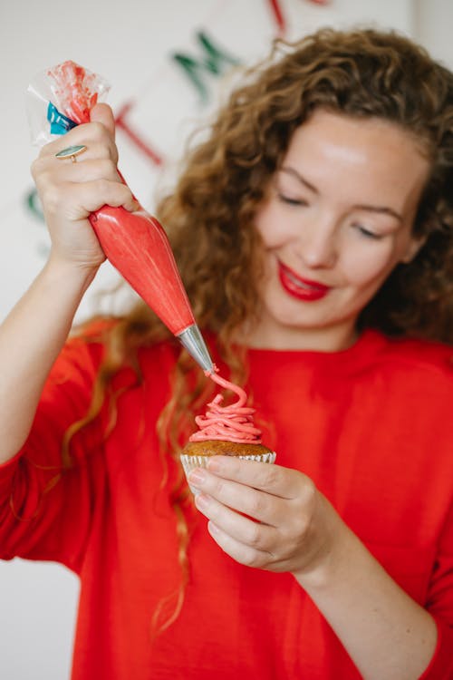 Cheerful female cook in red apparel and pastry bag decorating yummy cupcake with buttercream in house