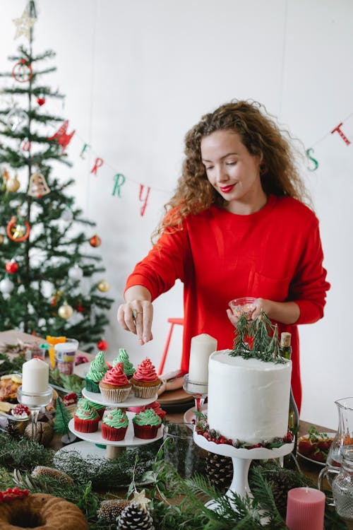 Smiling female cook sprinkling delicious cupcakes with buttercream on top while preparing for Christmas holiday at home