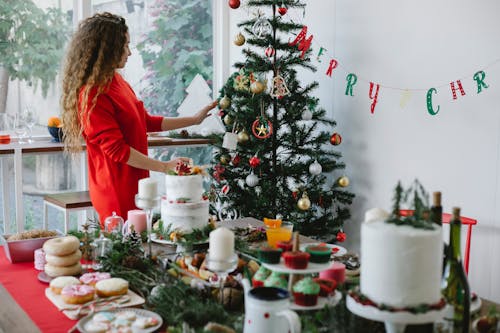 Woman decorating Christmas tree in house room with assorted desserts