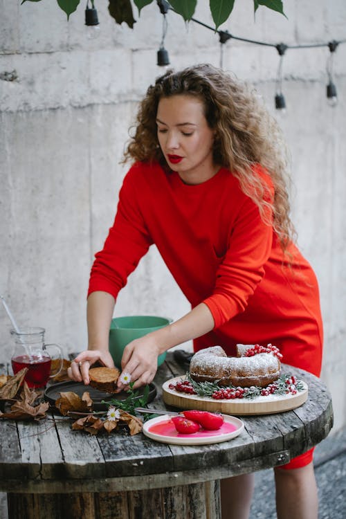 Adult female in red dress decorating wooden table with delicious cake during New Year holiday in patio