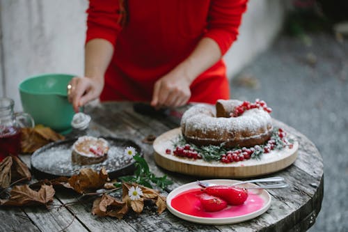 Crop anonymous female in red dress decorating freshly baked delicious pie slice with sugar powder on autumn backyard