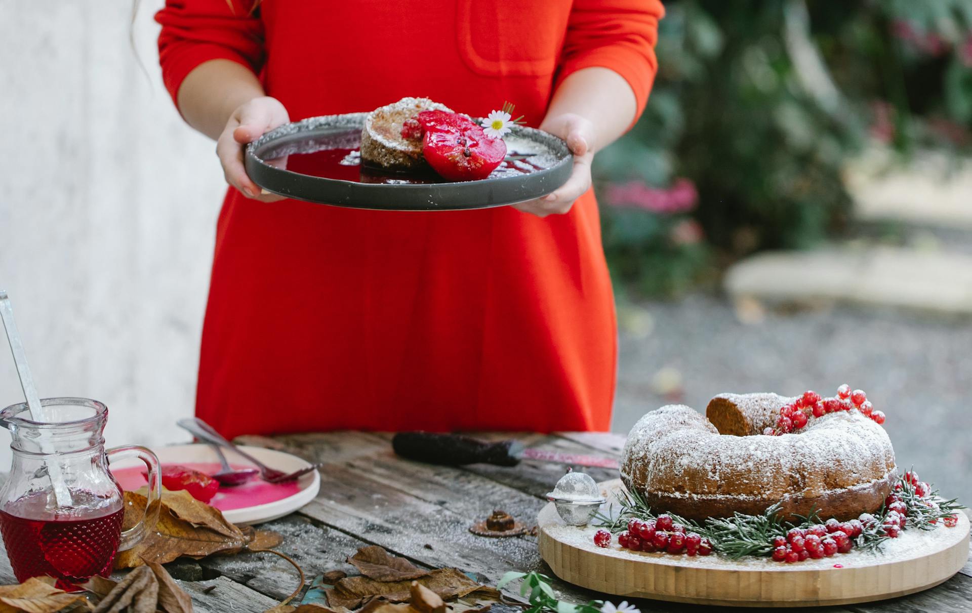 Crop unrecognizable female in red dress demonstrating plate with palatable homemade pie garnished with pear and chamomile flower while standing in backyard