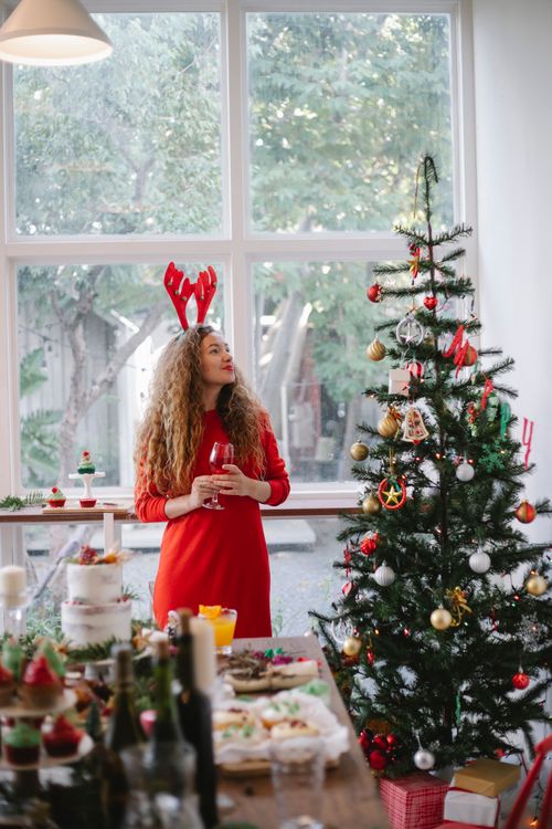 Free Cheerful female with long curly hair in bright clothes standing near Christmas tree in cozy room with festive decorations in daytime Stock Photo