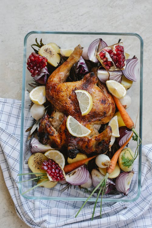 Free Overhead of tasty grilled whole chicken with lemon and pomegranate placed among onion with carrot served in glass bowl on towel Stock Photo
