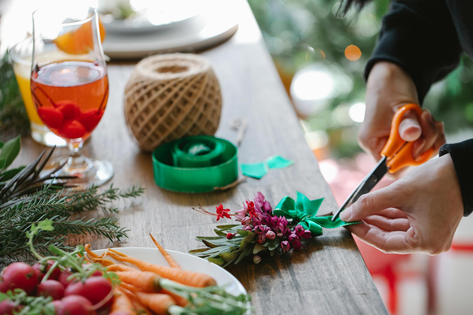 Unrecognizable male with scissors cutting ribbon of decorative green plant with small flowers while standing at table with vegetables and wineglass of drink on blurred background