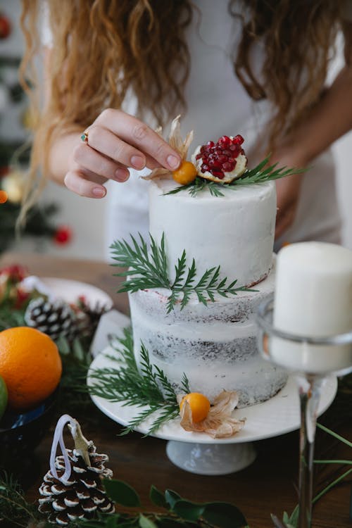 Crop anonymous female decorating homemade cake with fresh berries and herbs placed on table in exquisite plate for Christmas celebration