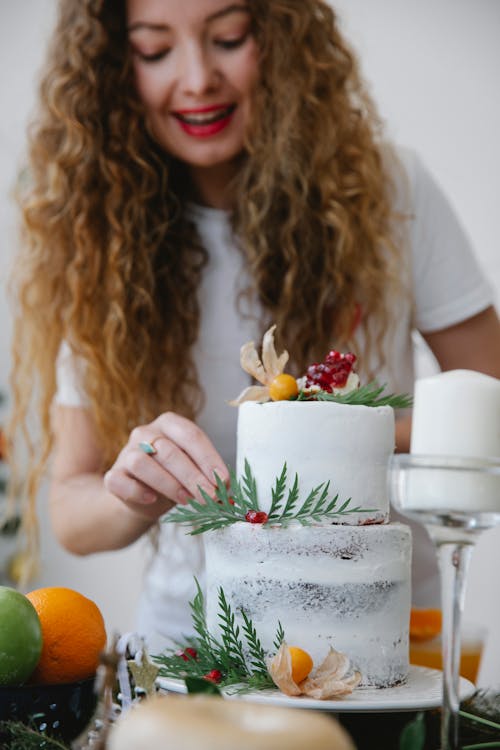 Free Young woman standing at table with assorted fruits and decorating cake with berries and herbs for Christmas party Stock Photo