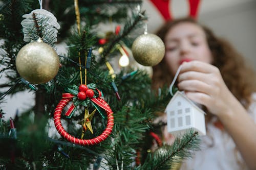 Crop anonymous female in warm sweater decorating Christmas tree with paper house bauble