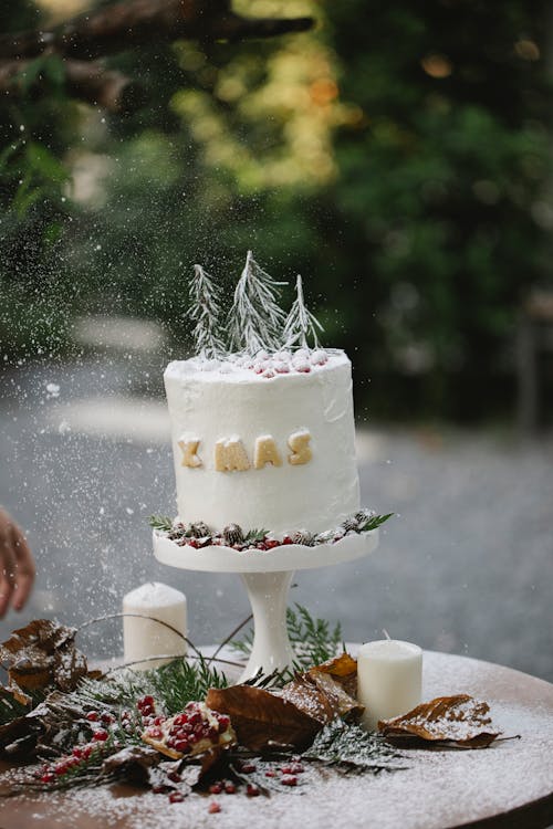 Christmas cake decorated with small Christmas trees on table in garden with dry leaves covered with snow and white candles