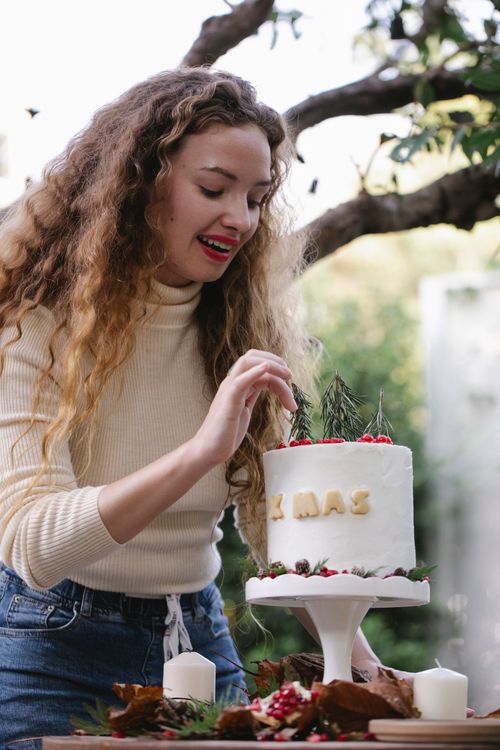 Free Low angle of attractive young female smiling and decorating delicious Christmas cake while standing at table in garden Stock Photo