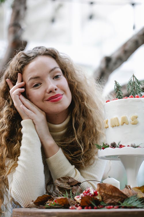 Smiling young woman touching cheek while sitting at table decorated with leaves near Christmas cake