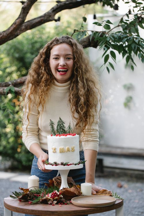Free Cheerful female cook looking at camera at table with delicious New Year cake with rosemary sprigs and berries on top Stock Photo