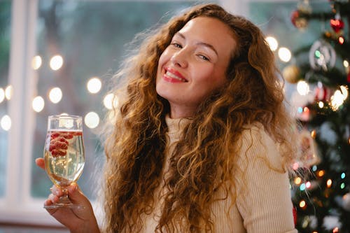 Content adult female with wavy hair and glass of alcoholic drink looking at camera during Christmas holiday at home