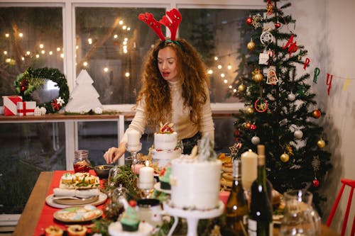 Adult female in decorative deer horns at table with assorted desserts preparing for New Year holiday in house