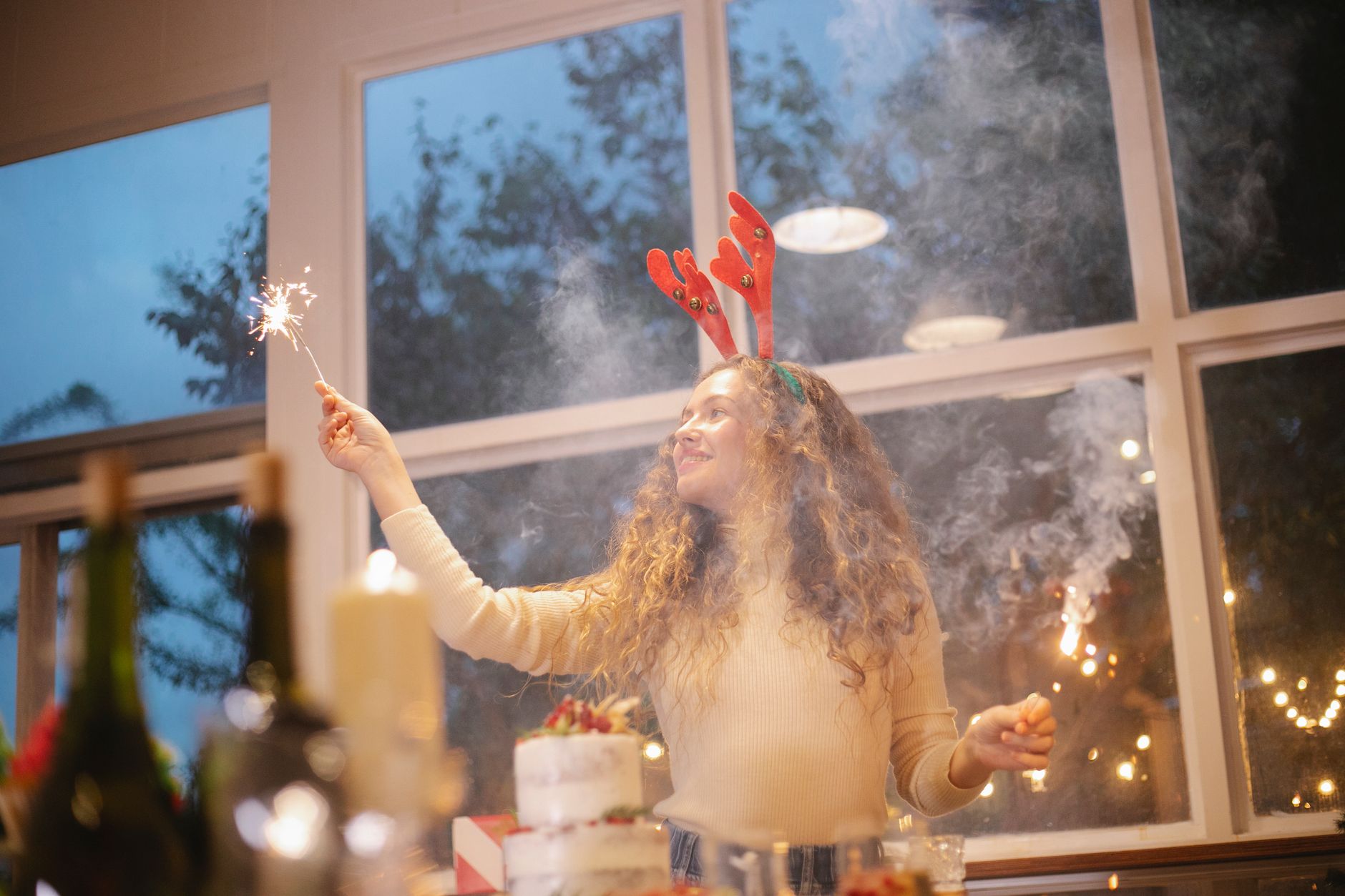 Content female chef in decorative deer horns with shiny sparkler celebrating New Year holiday while looking away in house