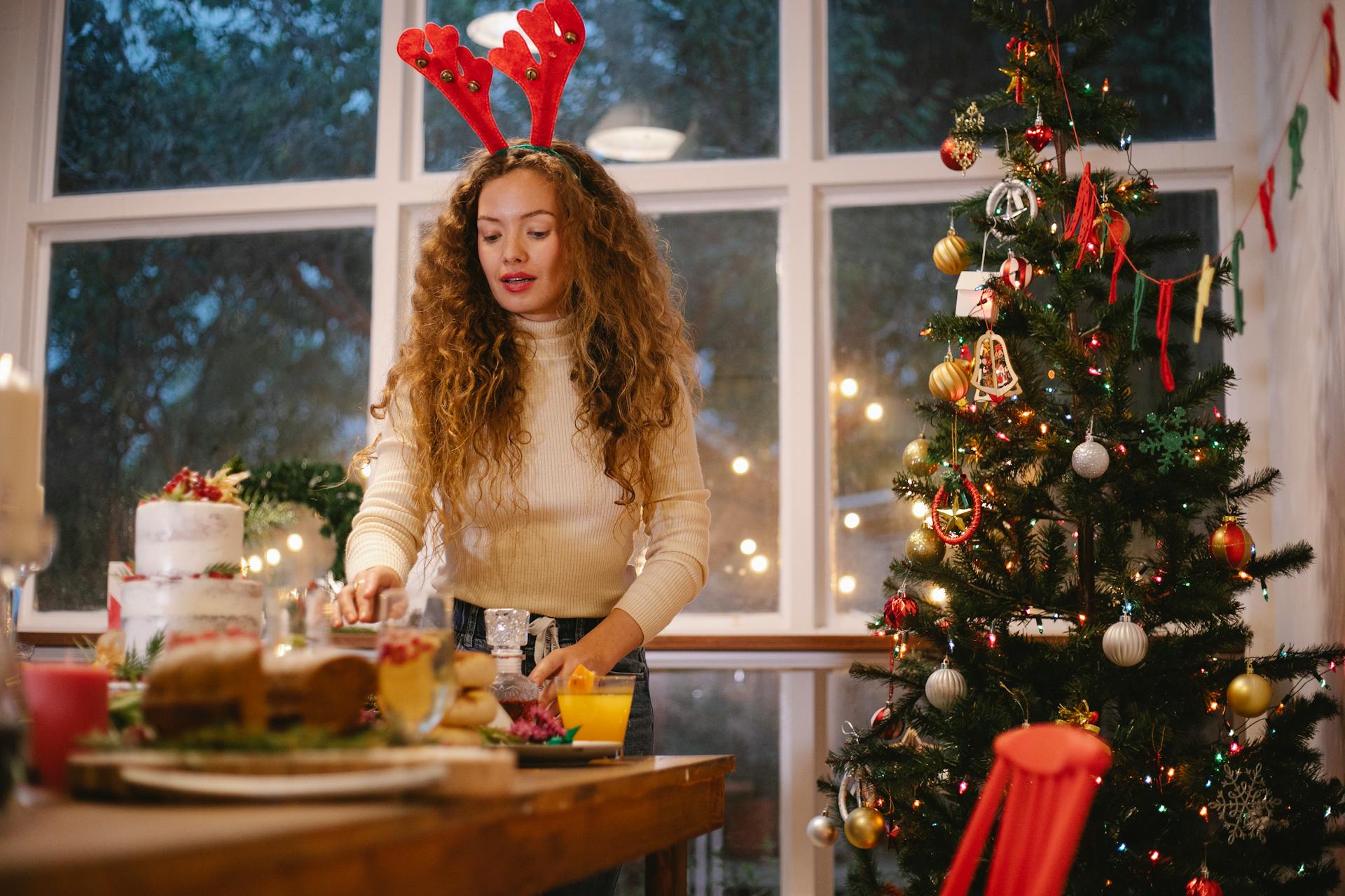 Adult female in decorative deer horns at table with delicious cake against fir tree during Christmas holiday in house