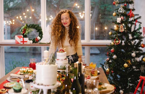 Sincere female in room with assorted desserts and fir tree with baubles against shiny lights during New Year holiday