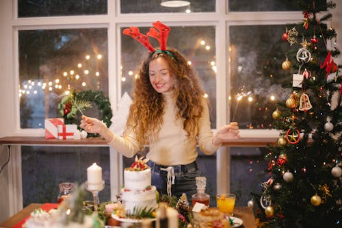 Cheerful female in decorative deer horns with burning sparklers standing at table with tasty cake for New Year holiday at home