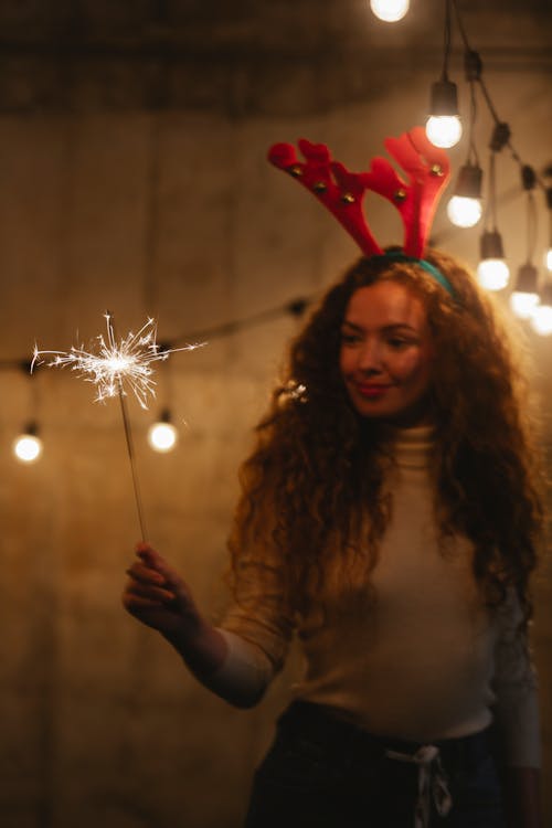 Friendly female in decorative deer horns with glowing sparkler celebrating New Year holiday under garland at dusk