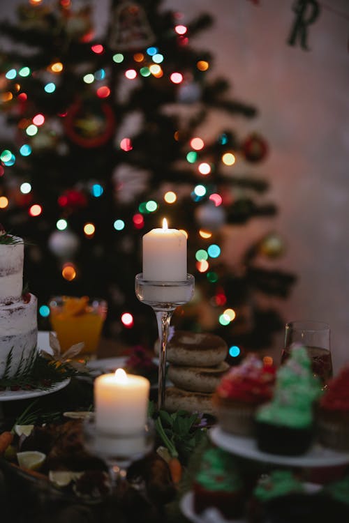 Flaming wax candles on table with assorted treats against fir tree with glowing lights during New Year holiday at home