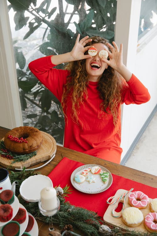 From above of cheerful female covering eyes with tasty gingerbread cookies at table with various treats during New Year holiday