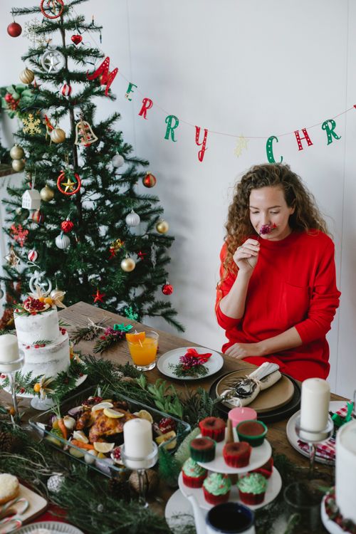 Free Romantic female enjoying scent of flower at table with assorted desserts against decorated fir tree during New Year holiday Stock Photo