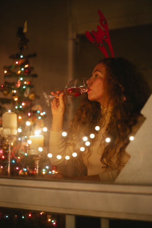 Through glass of female celebrating Christmas with glass of champagne in room with fir tree decorated with shiny garlands at night