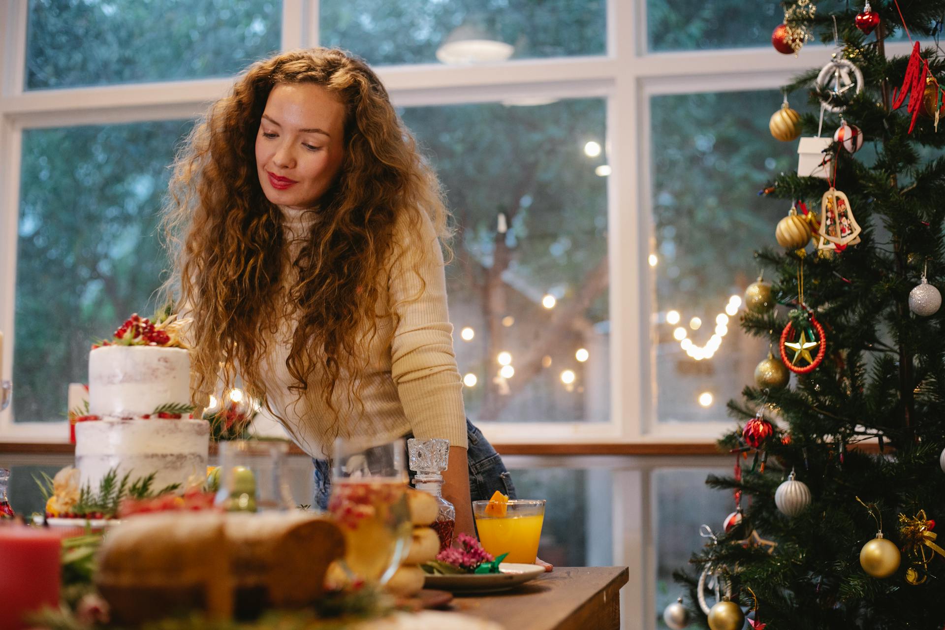 Low angle of serious female leaning on served festive table with two tier cake near decorated Christmas tree