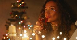 Through glass of dreamy female with glass of champagne sitting among shiny garlands and burning candles during Christmas celebration