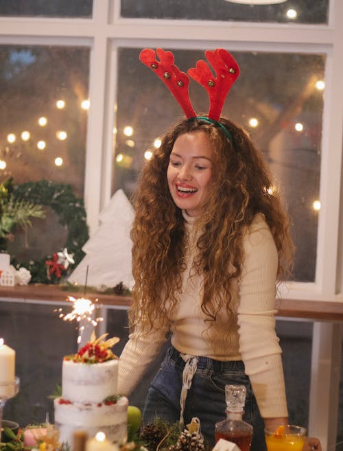 Delighted female in deer headband standing with shining Bengal light near table for Christmas celebration