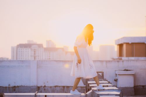 Free A woman in a white dress walks on a rooftop at sunset, with an urban cityscape in the background. Stock Photo