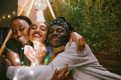 Free Three friends share a joyful embrace, radiating happiness under string lights. Stock Photo