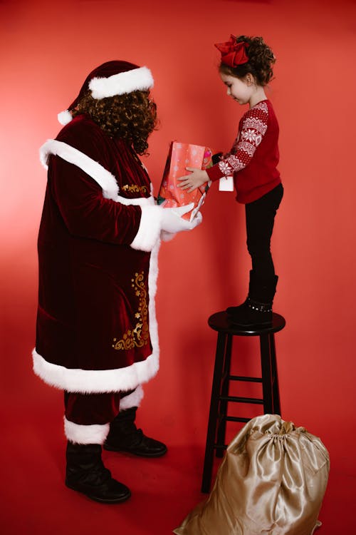 A Man in Santa Claus Costume Handing a Gift to a Girl Standing on a Bar Stool