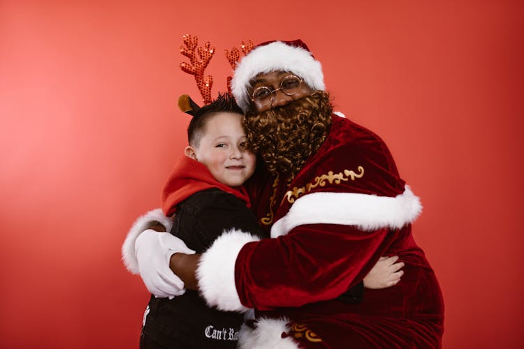 A Man In Santa Claus Costume Hugging A Boy In Black Hoodie