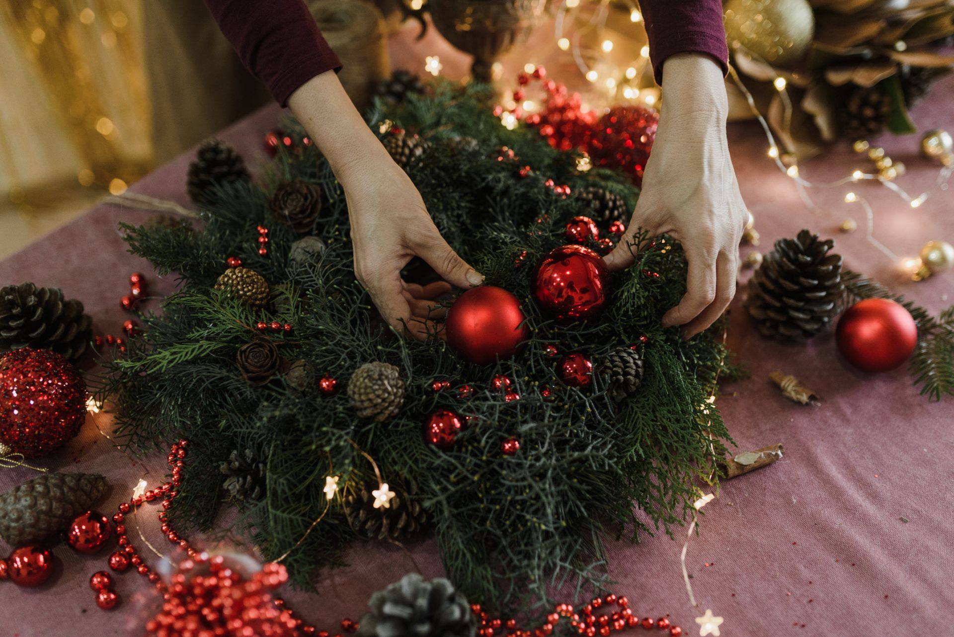 Hands decorating a festive Christmas wreath with red ornaments and pine cones on a table.