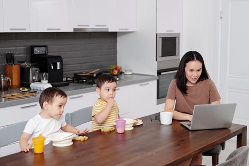 Free A mother multitasks on her laptop while her twin toddlers enjoy a snack in a modern kitchen setting. Stock Photo