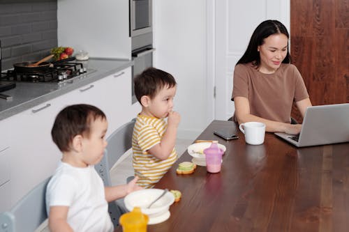 A Woman in Brown Shirt Working on Her Laptop while Sitting Near the Two Kids Eating Breakfast