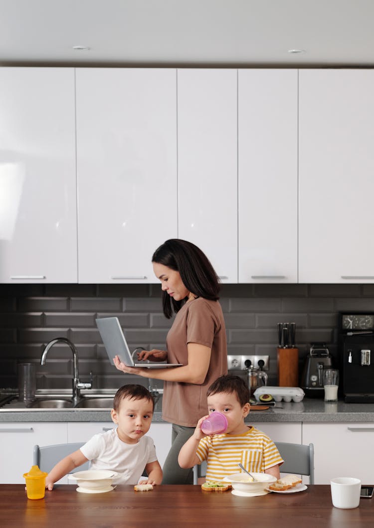 A Woman Working On Her Laptop While Standing Near Her Kids Eating Breakfast