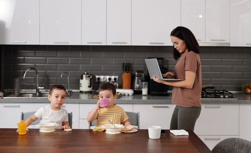 Free A mother multitasking with a laptop while her children enjoy breakfast at home. Stock Photo