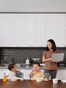 A mother balances work on her laptop while supervising her children during breakfast in a modern kitchen.