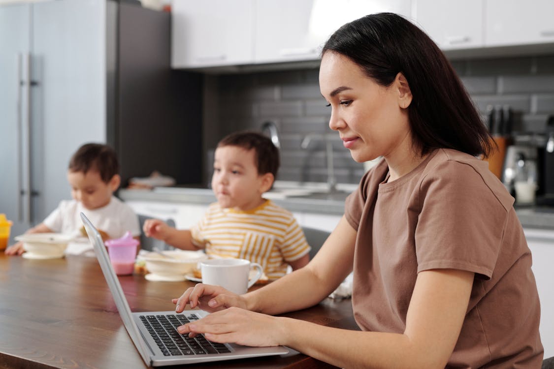 Free A Woman in Brown Shirt Typing on Her Laptop while Sitting Beside Her Kids Stock Photo