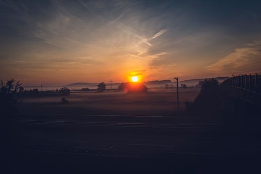 A tranquil sunset over foggy fields with a road and bridge silhouette, capturing nature's serene beauty.