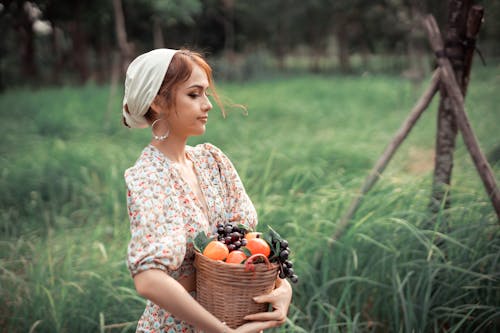 Side view of female farmer in summer clothes carrying basket with fresh fruits while standing on grassy field